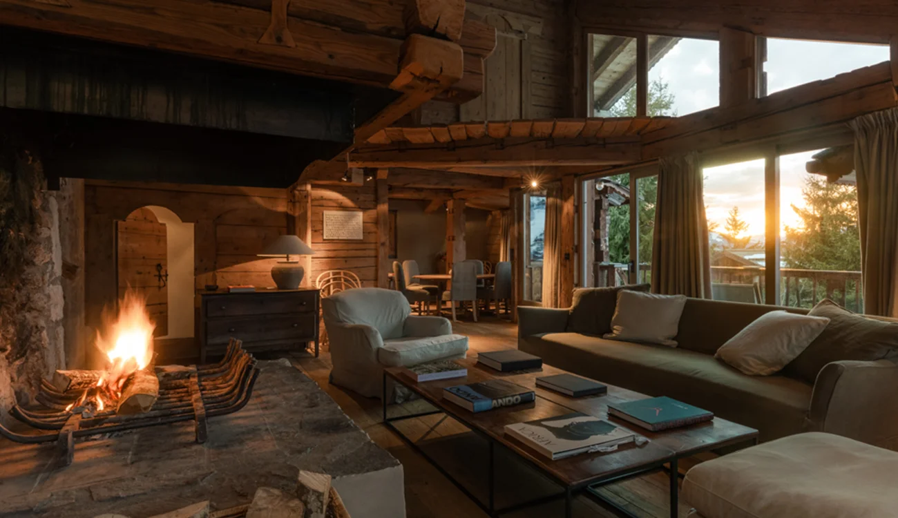A warmly lit Alpine chalet lounge bathed in golden sunset light, with mountain views through the window