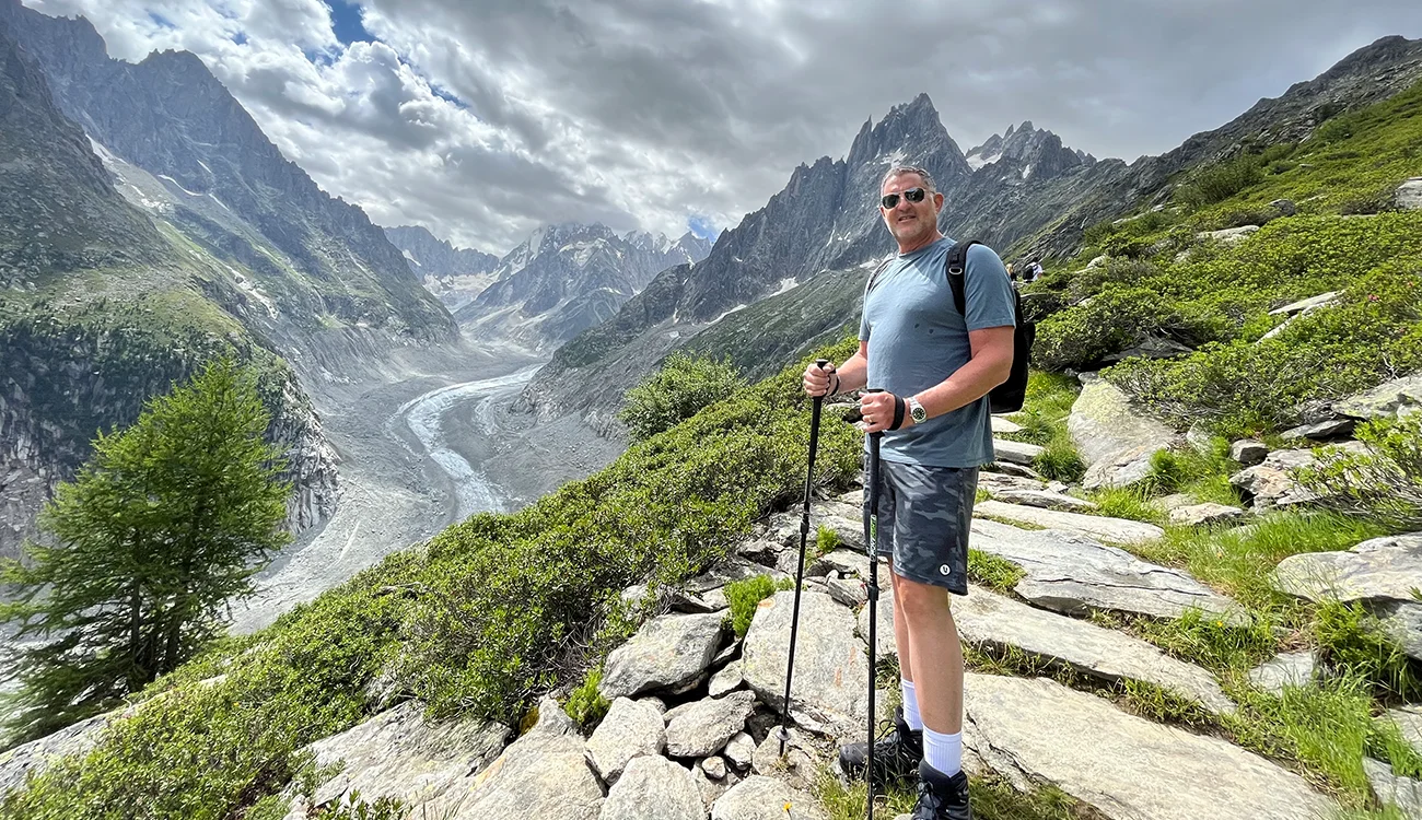 A hiker stands on a ridge overlooking a sweeping Alpine landscape of mountains and glacier below