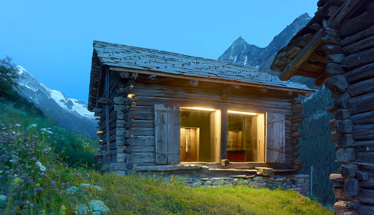 A luxury standalone mountain hut sits in isolation among dramatic Alpine peaks and meadows