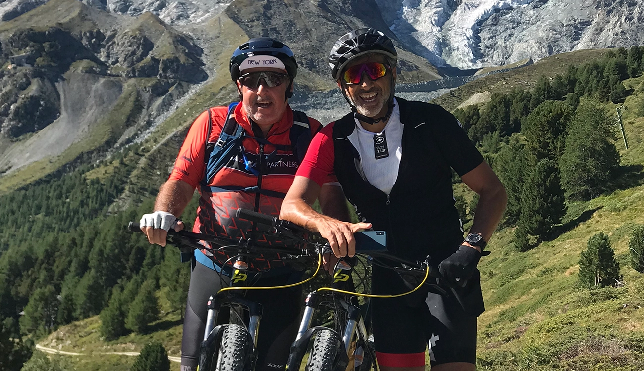 two-friends-e-bikes-alpine-mountains-swissskisafari-luxury-biking-safari-alps-1300x750 Two friends ride e-bikes together on an Alpine trail with dramatic mountain peaks surrounding them