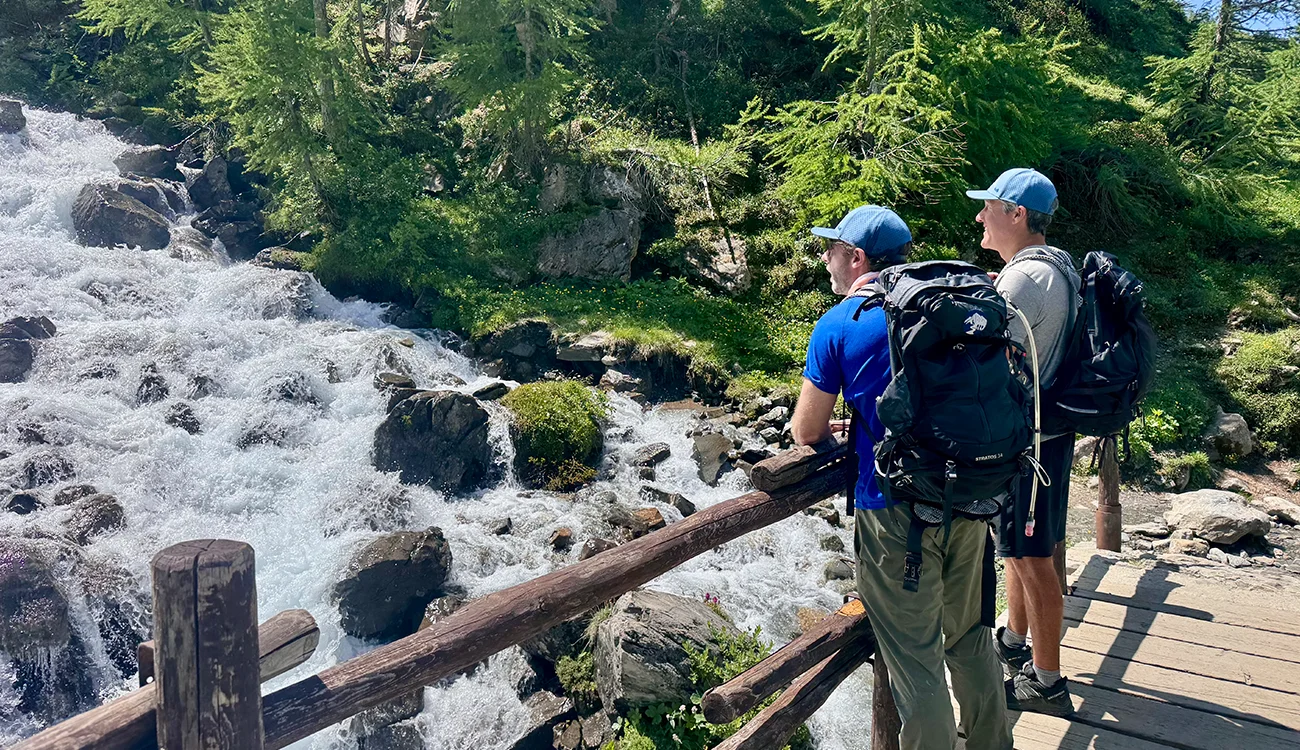 corporate-retreat-wooden-bridge-mountain-stream-alps Executives crossing a wooden footbridge above a fast-flowing mountain stream during a guided corporate hiking retreat in the Alps