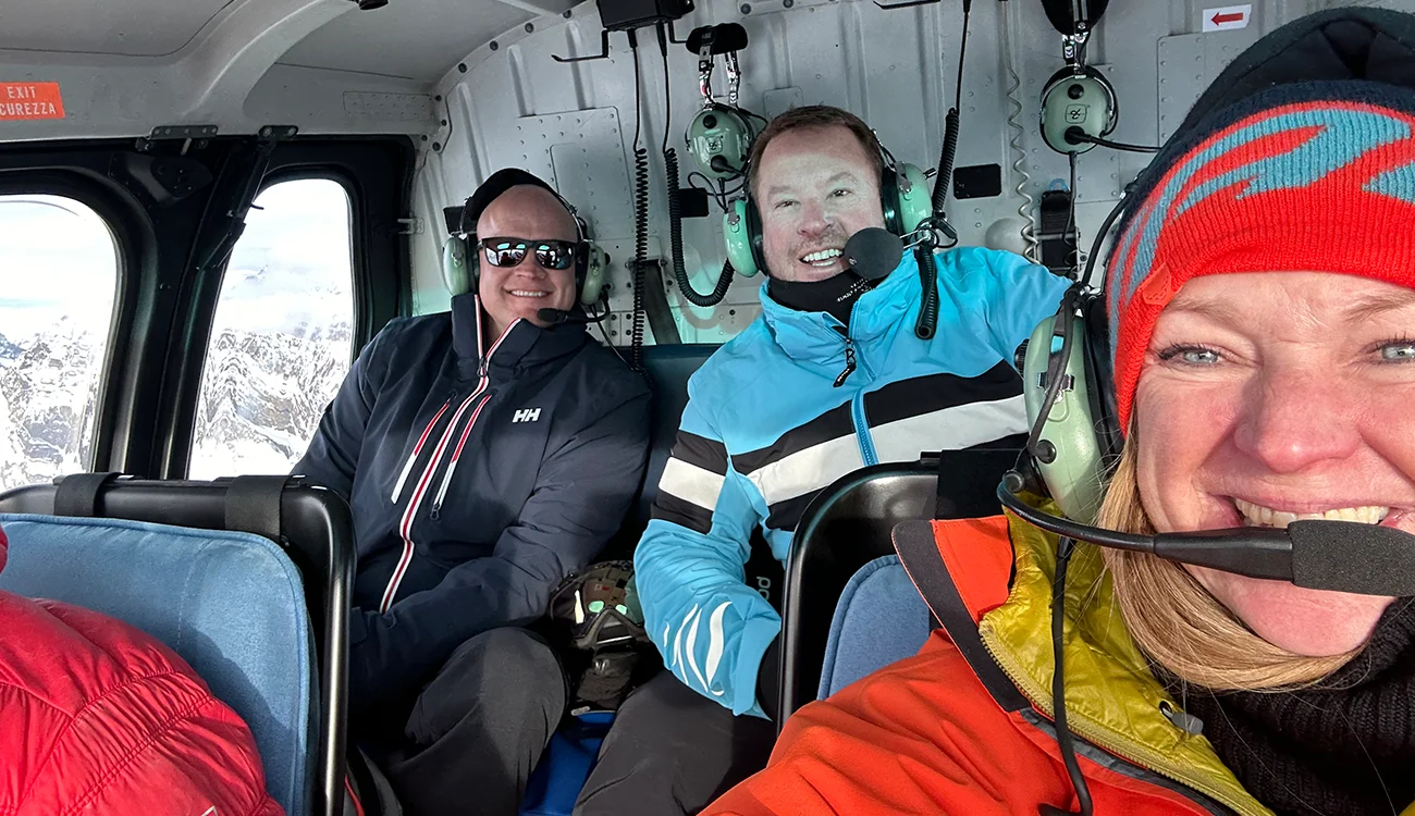 heli-ski-safari-guests-smiling-inside-helicopter Three smiling guests inside a helicopter during a SwisSkiSafari heli ski safari